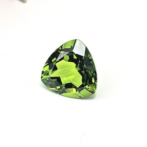 May include: A faceted, triangular-shaped, peridot gemstone. The gem is a vibrant, translucent green color with multiple facets that reflect light. The gemstone is set against a white background.