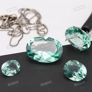 Op de afbeelding: Verschillende ovaalvormige, lichtblauwe edelstenen worden getoond op een zwart-wit oppervlak. Eén edelsteen wordt vastgehouden door een zilveren prong setting. Een zilveren ketting is zichtbaar op de achtergrond. De edelstenen zijn gefacetteerd.