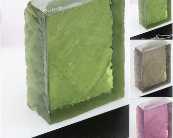 May include: A rectangular, translucent green gemstone pendant with a rough, textured surface. The pendant is encased in a clear, rectangular frame. Additional pendants in shades of green, brown, and purple are also shown.
