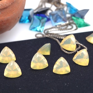 Op de afbeelding: Close-up van verschillende driehoekige, lichtgele edelstenen. Een zilveren ketting met een edelsteenhanger is zichtbaar, samen met andere blauwe en groene kristalachtige vormen. De edelstenen liggen op een zwarte ondergrond.