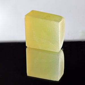 Puede incluir: Una barra de jab&oacute;n rectangular de color amarillo p&aacute;lido con una superficie lisa y ligeramente texturizada. El jab&oacute;n se refleja en una superficie negra.
