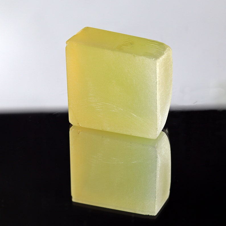 Puede incluir: Una barra de jab&oacute;n rectangular de color amarillo p&aacute;lido con una superficie lisa y ligeramente texturizada. El jab&oacute;n se refleja en una superficie negra.