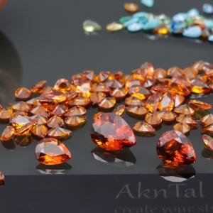 May include: A close-up of a collection of orange pear-shaped gemstones. The gemstones are scattered on a black surface. The gemstones are shiny and reflect light.