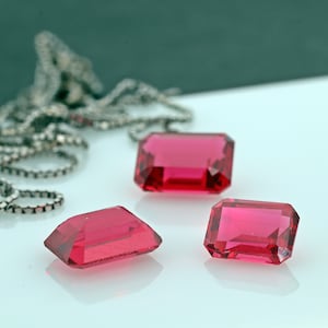 May include: Three red gemstone rectangular cut jewels on a white surface. The jewels are faceted and have a glossy finish.
