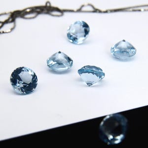 May include: Five light blue round faceted gemstones on a white surface. The gemstones are cut in a diamond shape.