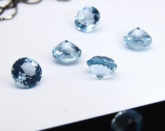 May include: Five round, faceted light blue gemstones are arranged on a white surface. Each stone features a brilliant cut with multiple triangular facets that reflect light. A thin, dark metallic chain is visible in the upper background.