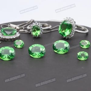 Op de afbeelding: Een assortiment groene edelstenen en sieraden. De afbeelding toont ovaal geslepen groene edelstenen, een hangerketting en twee ringen met een halo van kleinere stenen. De sieraden en edelstenen worden getoond op een donkere ondergrond.