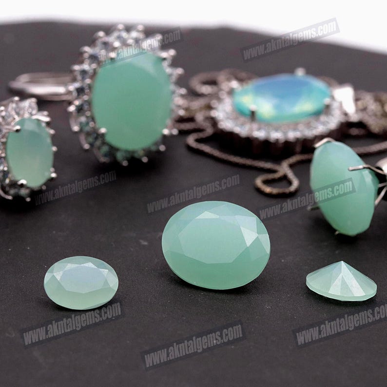 Op de afbeelding: Een assortiment aquamarijn edelstenen en sieraden. De afbeelding toont verschillende gefacetteerde, ovaalvormige edelstenen in verschillende maten, samen met ringen en een hanger. De edelstenen hebben een licht, zeegroene kleur en zijn geplaatst tegen een donkere achtergrond.