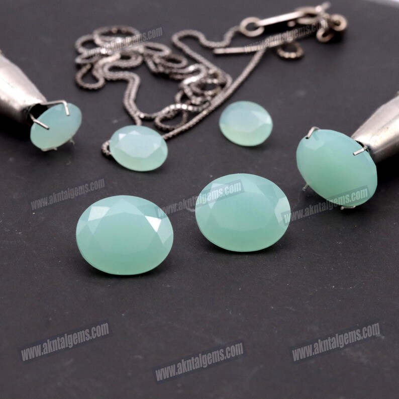 Op de afbeelding: Verschillende lichtblauwe-groene edelsteen cabochons in verschillende maten worden getoond op een donkere ondergrond. Een zilveren ketting en twee zilveren gereedschappen zijn ook aanwezig. De edelstenen hebben een facetgeslepen snede.