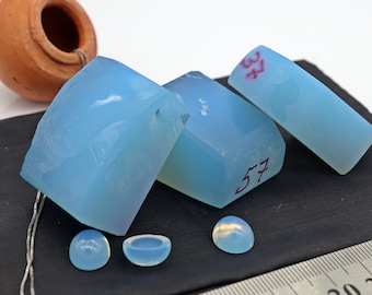 May include: A collection of light blue, translucent gemstones, including rectangular and rounded shapes, are displayed on a black surface. Some pieces have red numbers. A small, brown ceramic pot and a silver chain are also visible.
