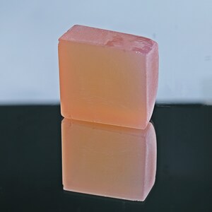 Puede incluir: Una barra de jab&oacute;n rectangular de color rosa claro y transl&uacute;cida. El jab&oacute;n est&aacute; sobre una superficie negra y se refleja en la superficie.