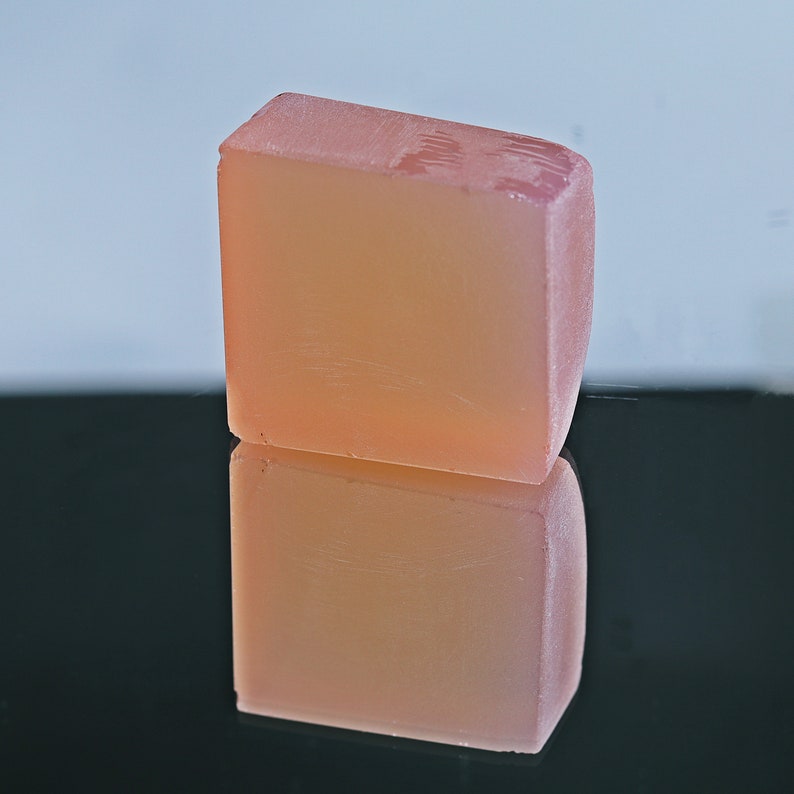 Puede incluir: Una barra de jab&oacute;n rectangular de color rosa claro y transl&uacute;cida. El jab&oacute;n est&aacute; sobre una superficie negra y se refleja en la superficie.