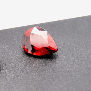 May include: A faceted, pear-shaped gemstone in a deep red colour. The gem has a polished surface with multiple facets that reflect light. The gemstone is set against a dark background, highlighting its colour and shape.