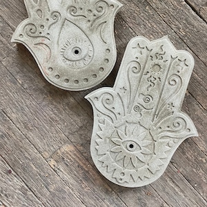 Op de afbeelding: Twee grijze betonnen hamsa hand wanddecoraties met ingewikkelde ontwerpen en een boze oog in het midden.