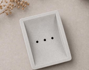 May include: A rectangular, light gray soap dish with three drainage holes. The dish has a slightly textured surface and a minimalist design. The soap dish is made of a solid material and is suitable for bathroom use.