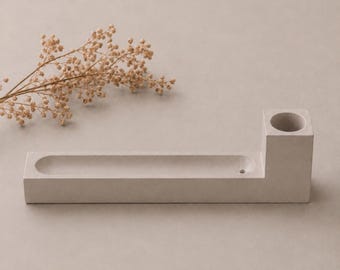 May include: A light grey concrete incense holder featuring a long, narrow rectangular tray with a rounded indentation and a raised square block on one end containing a circular opening for an incense stick, accompanied by a dried beige flower sprig.