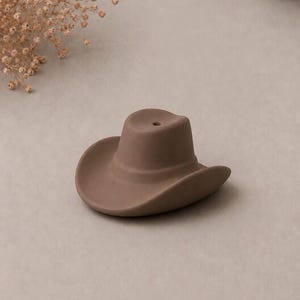 May include: A matte brown ceramic incense holder shaped like a cowboy hat. The hat features a curved brim and a central crown with a small circular hole at the top, designed to hold an incense stick securely in an upright position for burning.