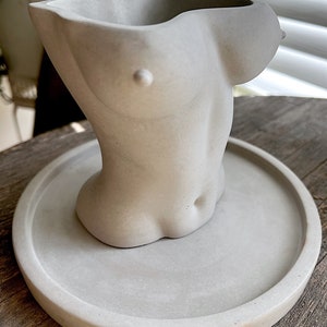 Może przedstawiać: Szara betonowa doniczka w kształcie torsu. Doniczka stoi na okrągłym, szarym betonowym podstawku.