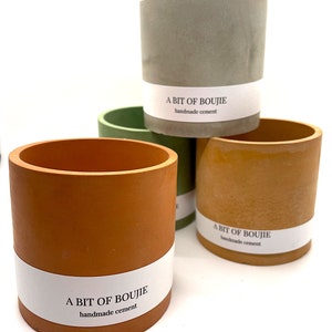 Könnte beinhalten: Drei Beton-Pflanztöpfe in verschiedenen Farben: grün, orange und braun. Jeder Topf hat ein weißes Etikett mit dem Text "A BIT OF BOUJIE handmade cement".