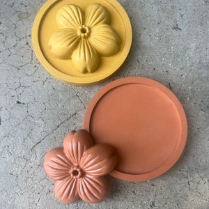 May include: Two round silicone moulds for making resin crafts. The larger mould is yellow and features a five-petal flower design. The smaller mould is brown and is plain.