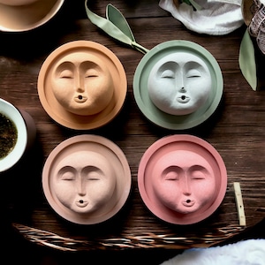 May include: Four round ceramic incense holders with a face design. Each holder is a different color: light brown, light green, light pink, and light beige. The faces have closed eyes and a small mouth.