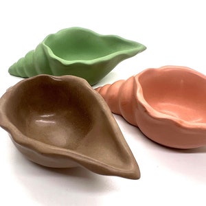 May include: Three ceramic seashell-shaped bowls in green, brown, and pink. The bowls are all in the shape of a seashell and have a smooth, glossy finish.