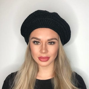 May include: A woman with long blonde hair is wearing a black beret. She is wearing a black shirt and has a neutral expression on her face.