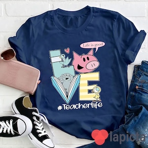 Op de afbeelding: Donkerblauw T-shirt met een afbeelding van een boek, een varken, een vogel en een eend. De afbeelding is in de vorm van het woord "LOVE". De tekst "Life is good" staat in een spreekballon boven het varken. De tekst "#Teacherlife" staat onder de afbeelding.