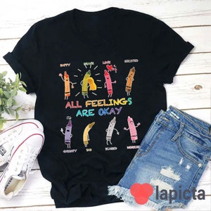 May include: Black t-shirt featuring a design of colourful crayons with faces and words such as "happy", "brave", "love", "excited", "grumpy", "sad", "scared", and "worried". The text "All Feelings Are Okay" is printed below the crayons.