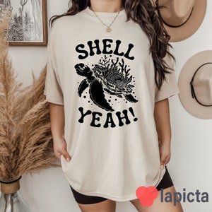 May include: A beige t-shirt with a black graphic of a turtle with coral and the text "SHELL YEAH!"