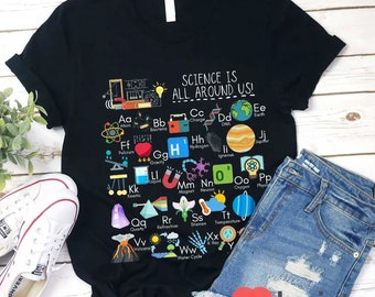 May include: A black t-shirt with a science-themed design. The design features a colorful alphabet with illustrations representing scientific concepts, such as atoms, bacteria, charge, DNA, earth, fulcrum, gravity, hydrogen, igneous rock, jupiter, kinetic energy, liquid, magnet, neuron, oxygen, physics, quartz, refraction, stamen, temperature, volcano, water cycle, and x-ray. The text "SCIENCE IS ALL AROUND US!" is printed at the top of the design.