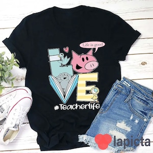 Op de afbeelding: Zwart T-shirt met een kleurrijke afbeelding van het woord "LOVE" gemaakt van een boek, een varken, een vogel en een eend. De tekst "Life is good" staat in een spreekballon boven het varken. De tekst "#Teacherlife" staat onder de afbeelding.