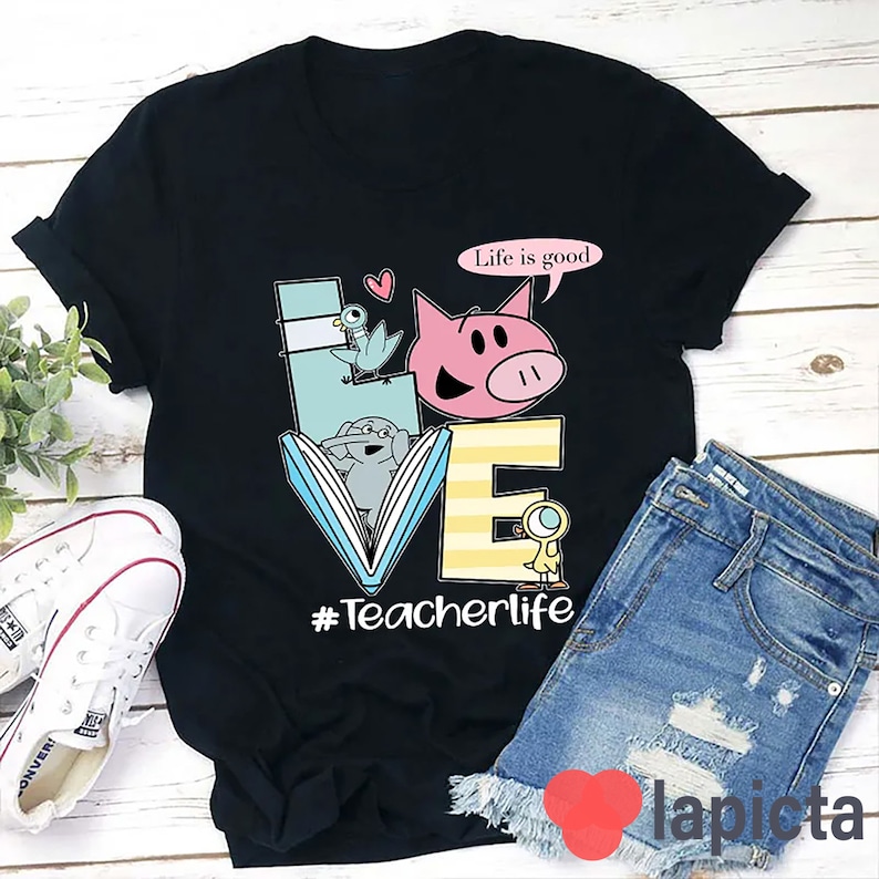 Op de afbeelding: Zwart T-shirt met een kleurrijke afbeelding van het woord "LOVE" gemaakt van een boek, een varken, een vogel en een eend. De tekst "Life is good" staat in een spreekballon boven het varken. De tekst "#Teacherlife" staat onder de afbeelding.