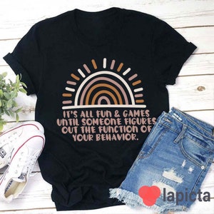 May include: Black t-shirt featuring a rainbow graphic and the text "It's all fun & games until someone figures out the function of your behaviour."