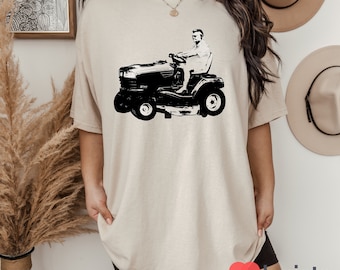 May include: A beige t-shirt with a black and white graphic of a person driving a lawnmower.