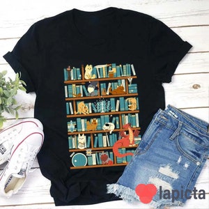 May include: Black t-shirt with a graphic of a bookshelf filled with books and various animals, including cats, a dragon, and a bird. The bookshelf is brown with white shelves and the books are blue, green, and red.