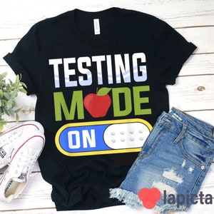 May include: Black t-shirt with the text "Testing Mode On" in white letters. The text is styled to look like a light switch with a red apple in the middle of the word "Mode".