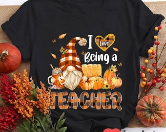May include: Black t-shirt featuring a gnome wearing a plaid hat and holding a cup of coffee. The text "I Love Being a Teacher" is written in orange and black plaid font with a heart that says "Love" in the middle.