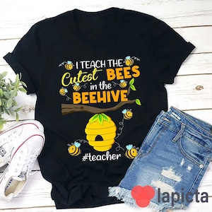 以下が含まれることがあります： 「I teach the Cutest BEES in the BEEHIVE」という文字が黄色と白でプリントされた黒いTシャツ。漫画のミツバチと黄色の蜂の巣のデザイン。蜂の巣の下には「#teacher」の文字。