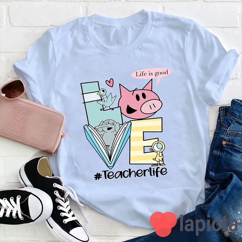 Op de afbeelding: Lichtblauw T-shirt met een afbeelding van een varken, een vogel en een olifant die een boek lezen. De afbeelding is omringd door de letters "LOVE" en de tekst "Life is good" en "#Teacherlife".