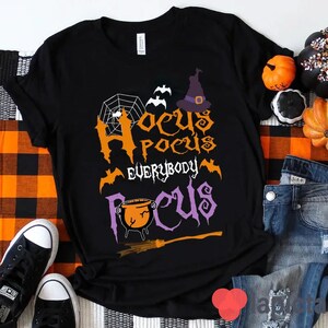 Puede incluir: Camiseta negra con un gráfico de temática de Halloween que dice "Hocus Pocus Everybody Focus" con un sombrero de bruja, murciélagos, una telaraña y un caldero.