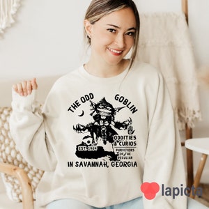 May include: A white sweatshirt with a black graphic of a goblin holding a sign that says "The Odd Goblin, Oddities & Curios, Est. 1984, Purveyors of the Peculiar, In Savannah, Georgia".