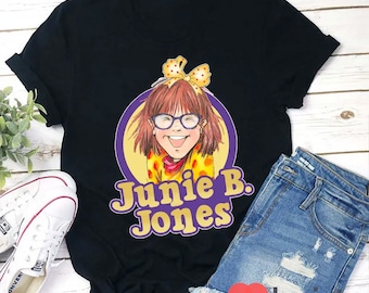 Op de afbeelding: Zwart T-shirt met een cartoonillustratie van Junie B. Jones die een geel en oranje gestreept shirt en een rode strik draagt. De tekst "Junie B. Jones" staat in paarse en gele letters.