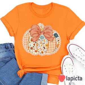May include: Orange t-shirt featuring a pumpkin design with a gingham bow and floral details. The pumpkin has a lace trim. Paired with blue jeans and white sneakers.