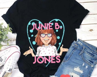 Op de afbeelding: Zwart T-shirt met een cartoonillustratie van Junie B. Jones in een roze hart met blauwe stippen. De tekst "Junie B. Jones" is roze.