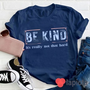 Puede incluir: Una camiseta azul marino con el texto "BE KIND it's really not that hard" en una fuente blanca desgastada con un contorno rosa.