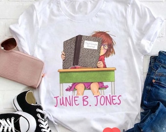 Op de afbeelding: Wit T-shirt met een cartoonillustratie van een meisje dat aan een bureau zit en een boek leest waarop "First Grade Journal" staat. De tekst "Junie B. Jones" staat onder de illustratie.