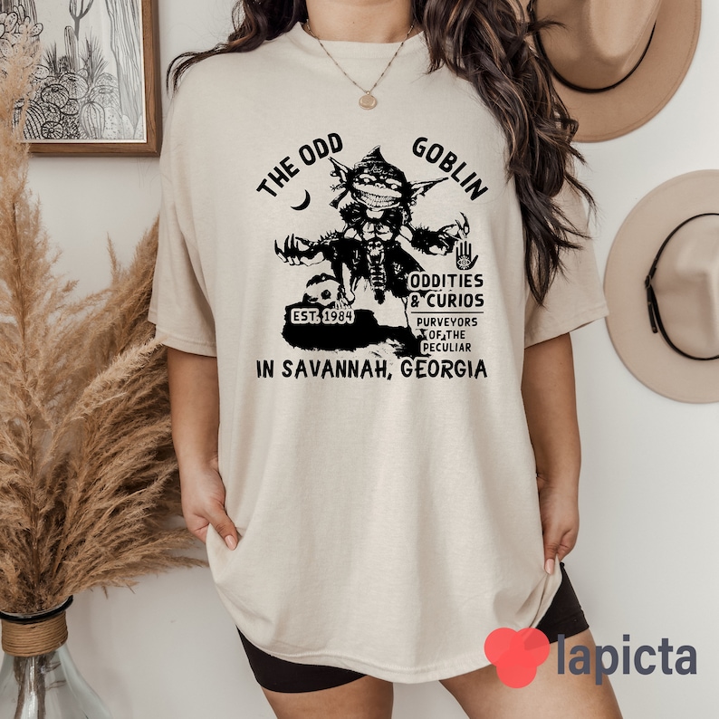 May include: A beige t-shirt with a black graphic of a goblin holding a skull. The graphic includes the text "The Odd Goblin", "Est. 1984", "Oddities & Curios", "Purveyors of the Peculiar", and "In Savannah, Georgia".