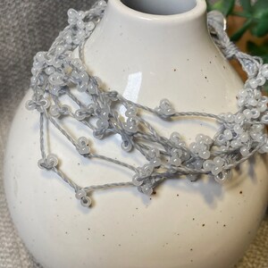 May include: A delicate, handmade necklace featuring strands of gray thread adorned with small, light gray beads. The necklace is draped around a white ceramic vase with a speckled finish, creating a soft, neutral color palette. The beads are clustered to resemble tiny flowers.
