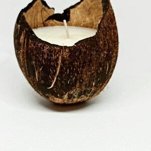 Può includere: Una candela in un guscio di cocco naturale. La candela è riempita di cera bianca e ha uno stoppino. Il guscio di cocco è marrone e ha una superficie strutturata, su sfondo bianco.